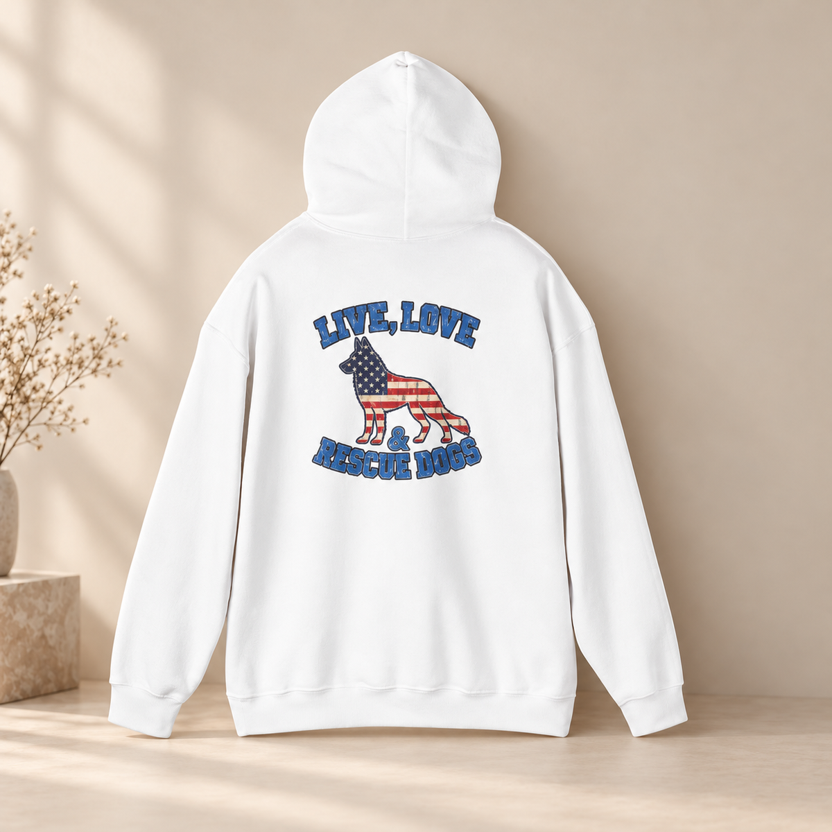 A white pullover hoodie shown from the back, featuring a graphic of a dog filled with an American flag pattern and the words 'LIVE, LOVE & RESCUE DOGS' in blue. The hoodie is displayed on a light wooden surface against a warm beige background with soft shadows and a small vase of dried flowers on the left.