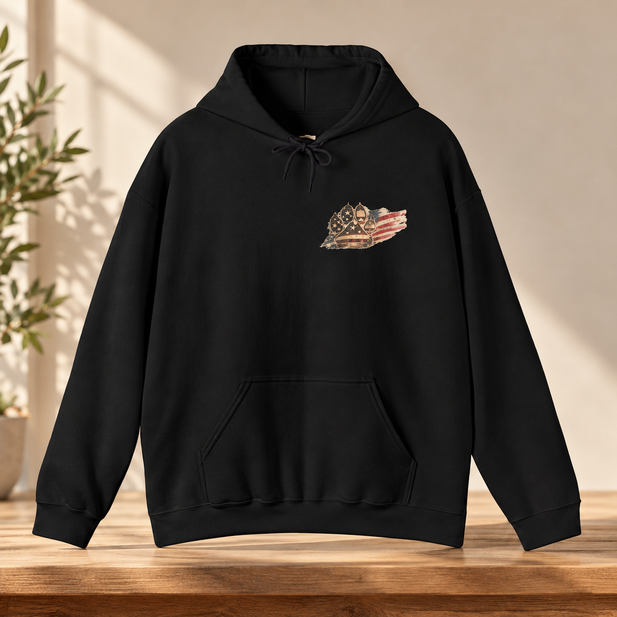 A black pullover hoodie with a small patriotic chest graphic, displayed on a wooden table in a warm, sunlit room setting.