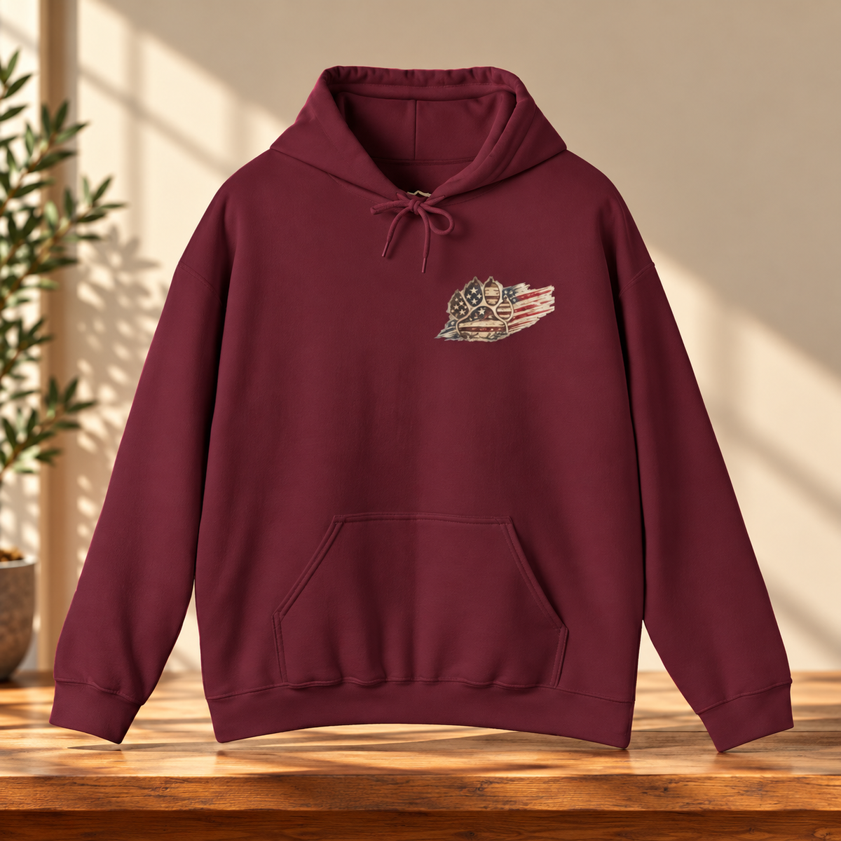 A burgundy pullover hoodie with a small distressed American flag and star graphic on the left chest, displayed on a wooden table with warm sunlight and shadows.