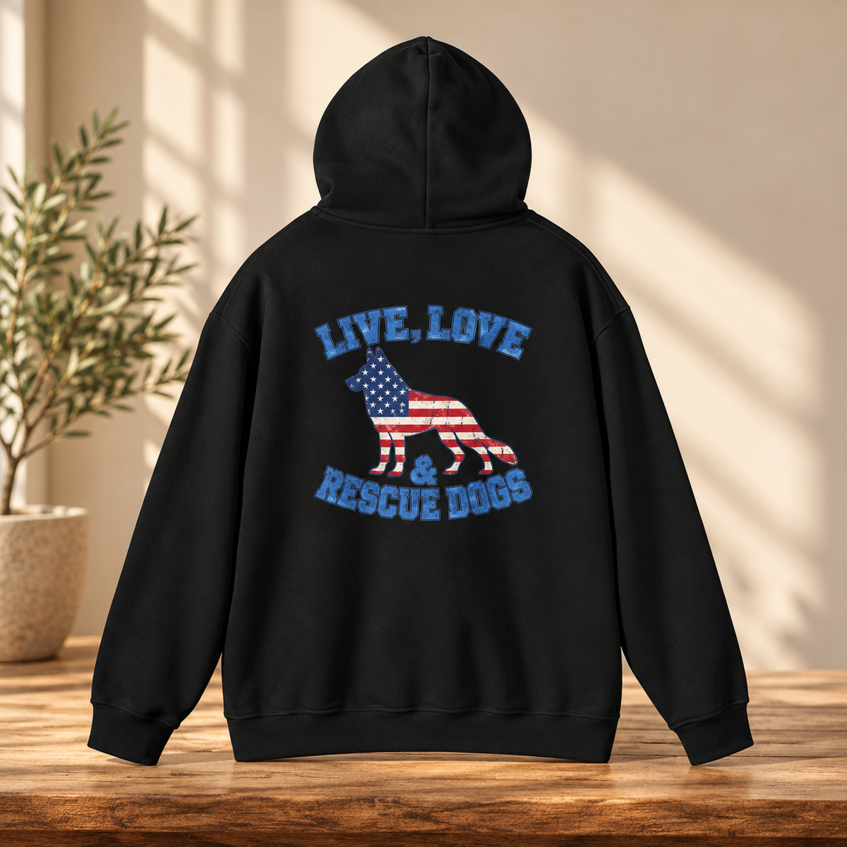 A black pullover hoodie shown from the back, placed on a wooden table in a sunlit room. The back features a large graphic with the words “LIVE, LOVE & RESCUE DOGS” in blue around a dog silhouette filled with an American flag pattern in red, white, and blue.