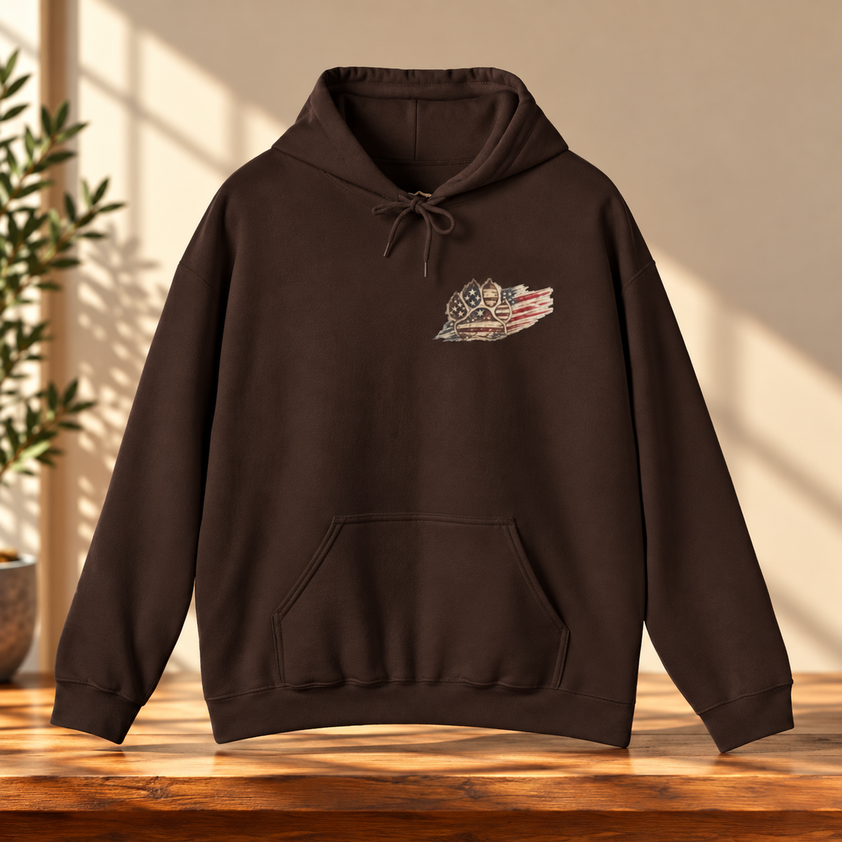 A dark brown pullover hoodie with a small patriotic chest graphic, displayed on a wooden table with warm sunlight and shadow patterns.