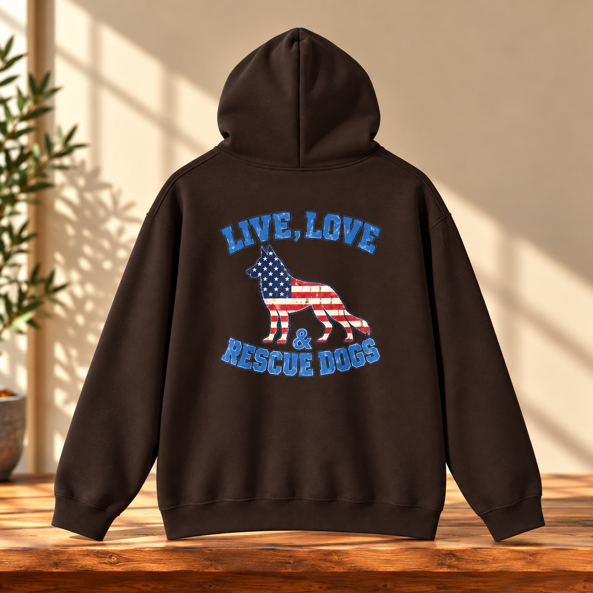 A dark brown hoodie shown from the back, featuring a graphic of a dog filled with an American flag pattern and the words 'LIVE, LOVE & RESCUE DOGS' in blue. The hoodie is displayed on a wooden table with a warm beige wall and a plant in the background.