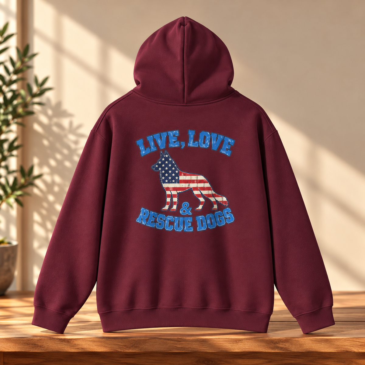 A maroon hoodie shown from the back, featuring a graphic of a dog filled with the American flag and the text 'LIVE, LOVE & RESCUE DOGS' in blue. The hoodie is displayed on a wooden surface with a warm beige background and soft window shadows.
