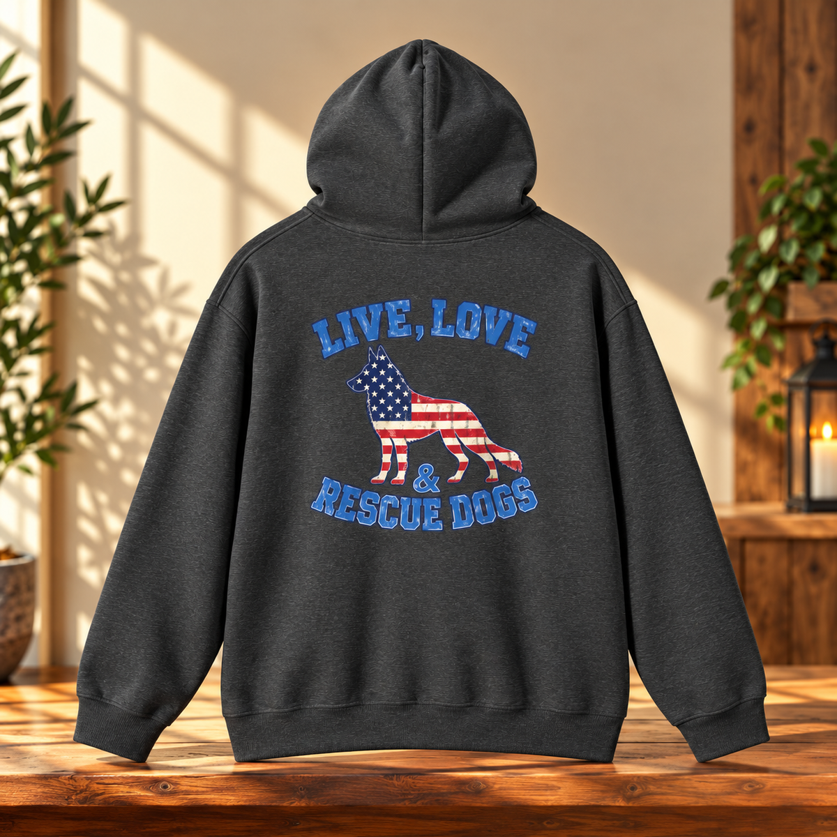 A dark charcoal gray hoodie shown from the back, featuring a graphic of a dog filled with the American flag and the text 'LIVE, LOVE & RESCUE DOGS' in blue. The hoodie is displayed on a wooden table in a warm, rustic room setting with plants and a lantern in the background.