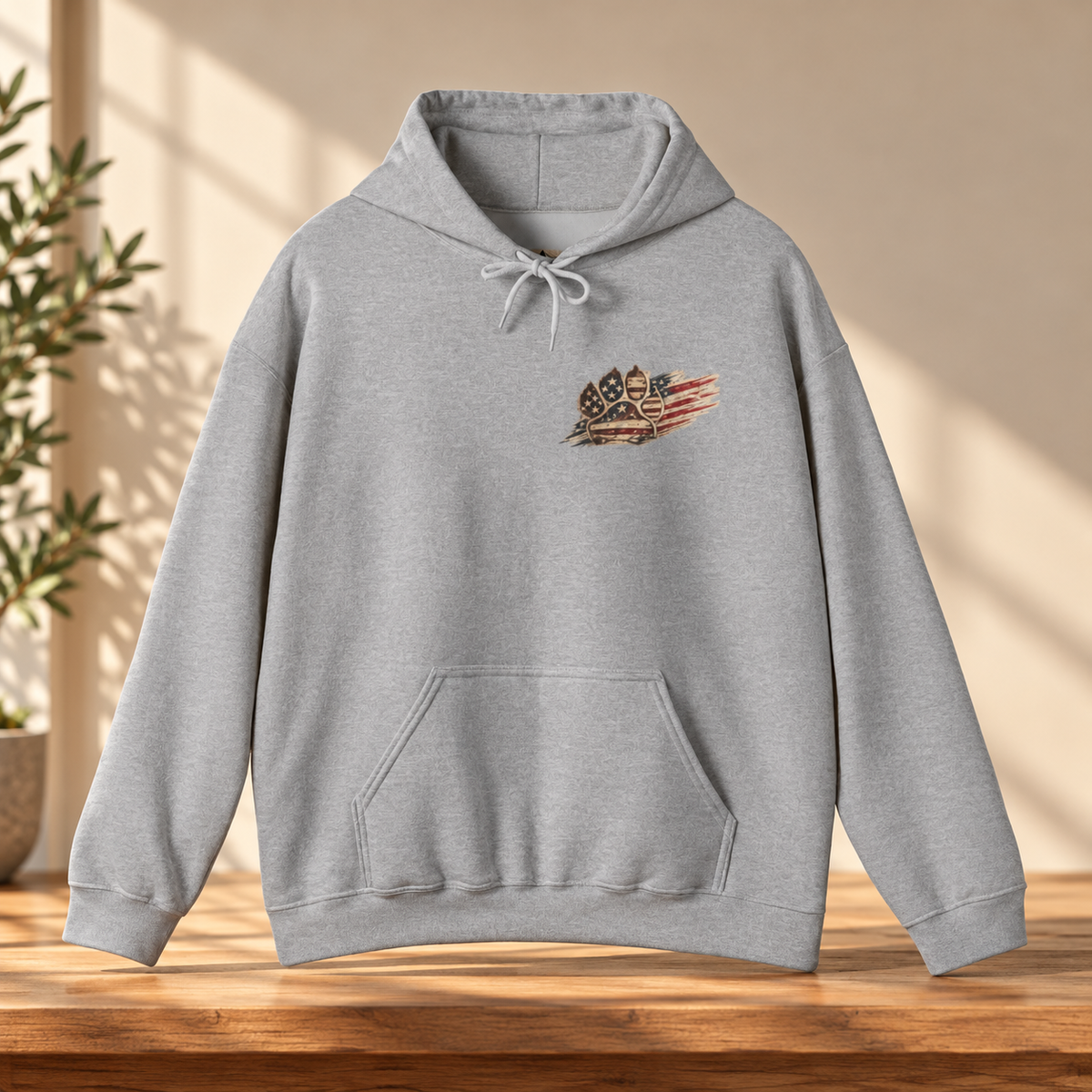 A light gray heather pullover hoodie with a small patriotic chest graphic, displayed on a wooden table with warm sunlight and a plant in the background.