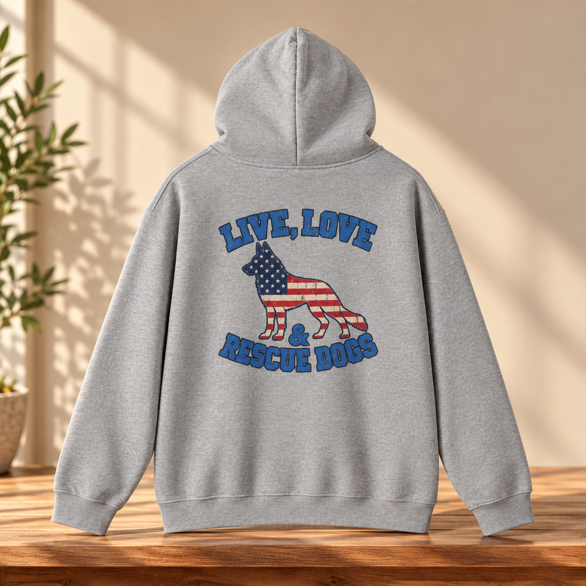 A light gray heather hoodie shown from the back, featuring a graphic of a dog filled with the American flag and the words “LIVE, LOVE & RESCUE DOGS” in blue. The hoodie is displayed on a wooden table with a warm beige wall and soft window shadows in the background.