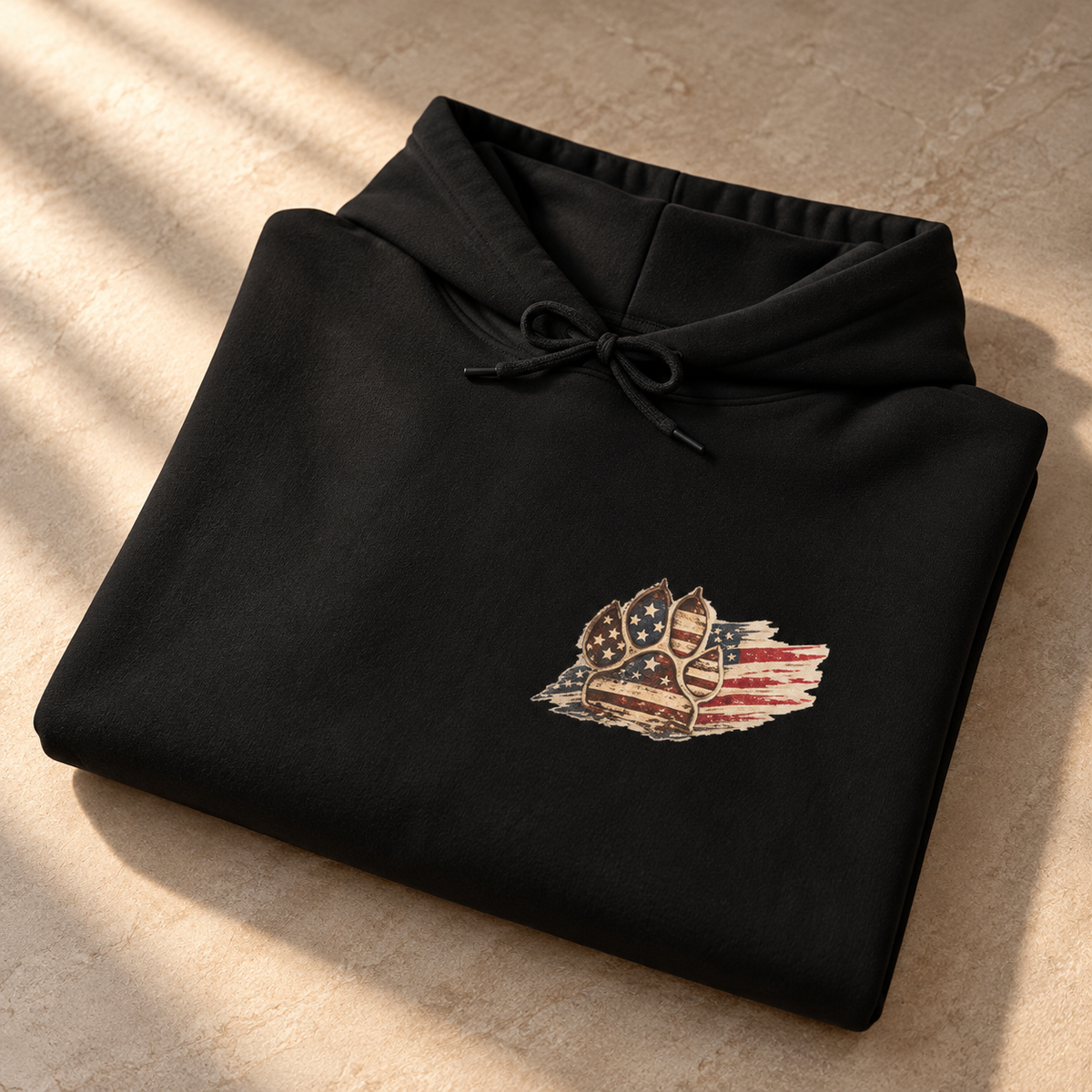 A folded black hoodie with a distressed American flag and shield graphic on the chest, displayed on a textured beige stone surface with soft diagonal shadows.