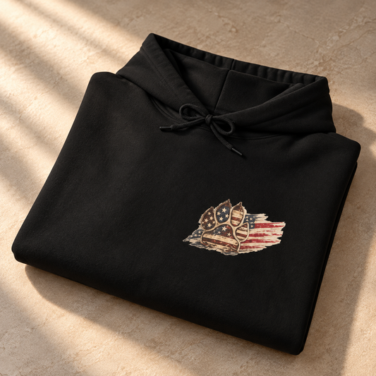 A folded black hoodie with a distressed American flag and shield graphic on the chest, displayed on a textured beige stone surface with soft diagonal shadows.