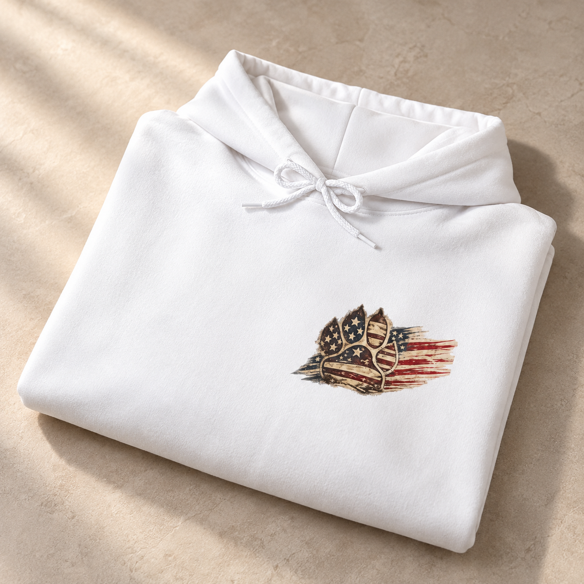 A folded white hoodie with a small chest graphic showing a paw print over a distressed American flag, placed on a beige textured surface with soft diagonal shadows.