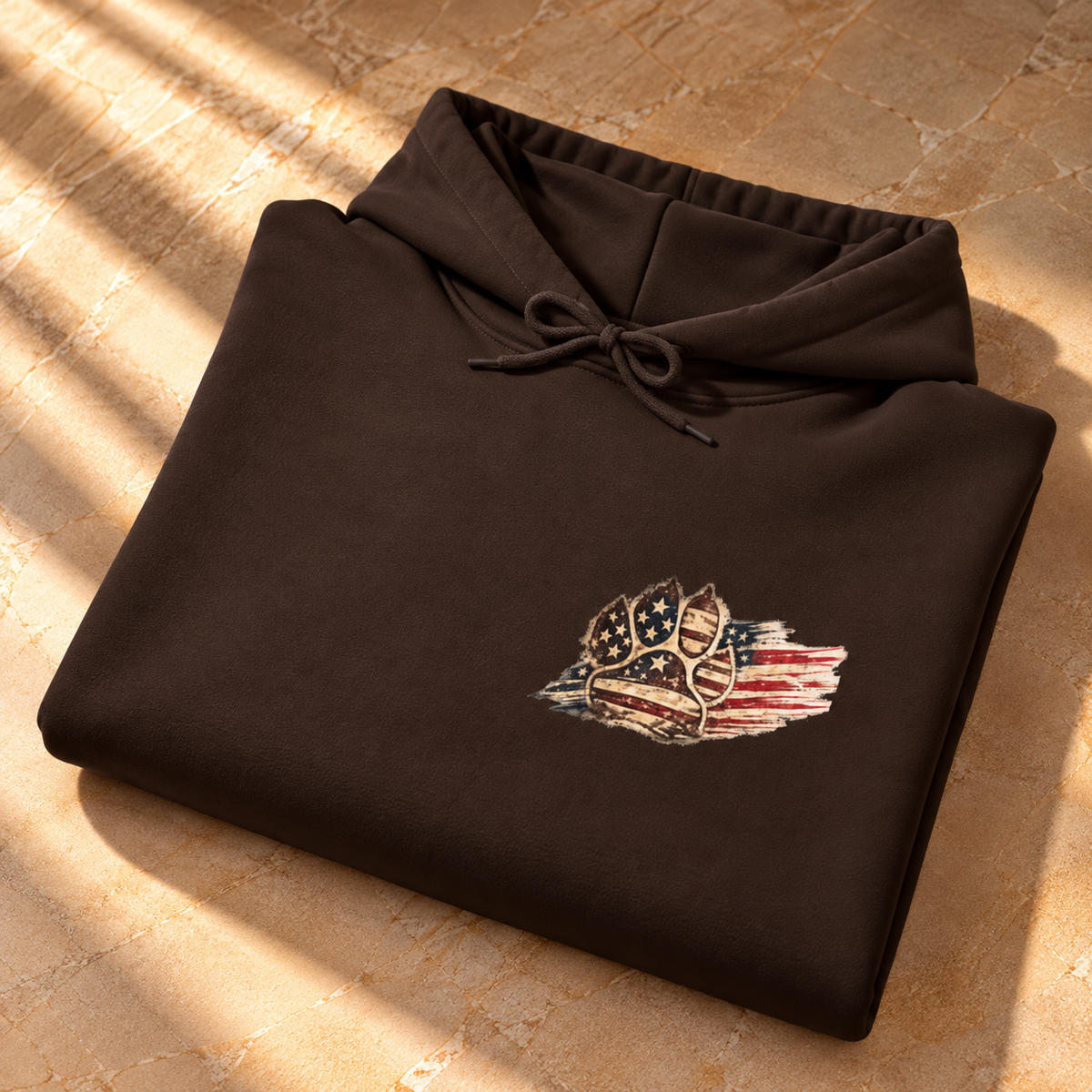 A folded dark brown hoodie with a drawstring hood, featuring a distressed American flag and shield graphic on the left chest, displayed on a textured stone surface with warm sunlight and shadows.