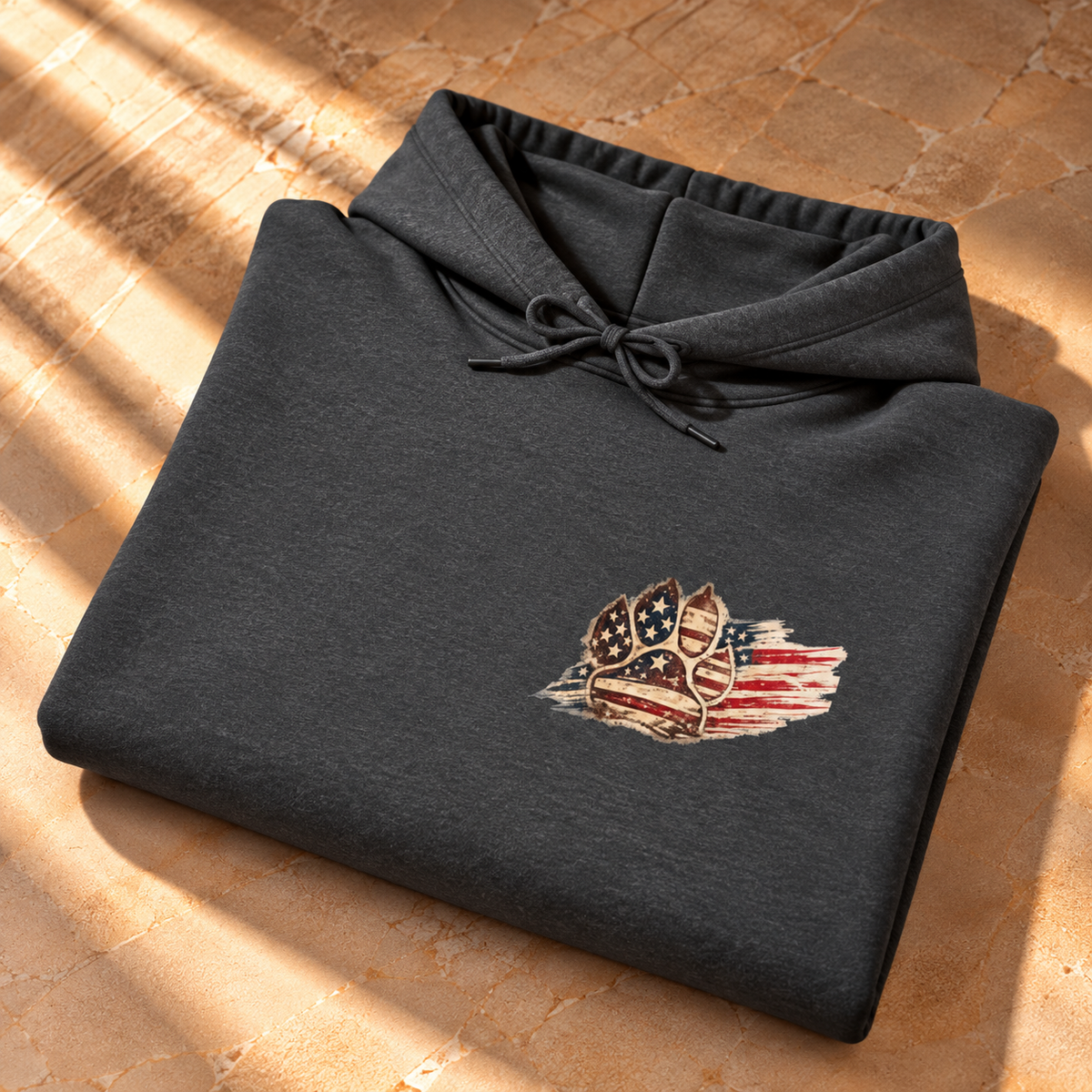 A folded dark charcoal gray hoodie with a drawstring hood, featuring a distressed American flag graphic with a shield emblem on the left chest, displayed on a textured stone surface with warm sunlight and shadows.