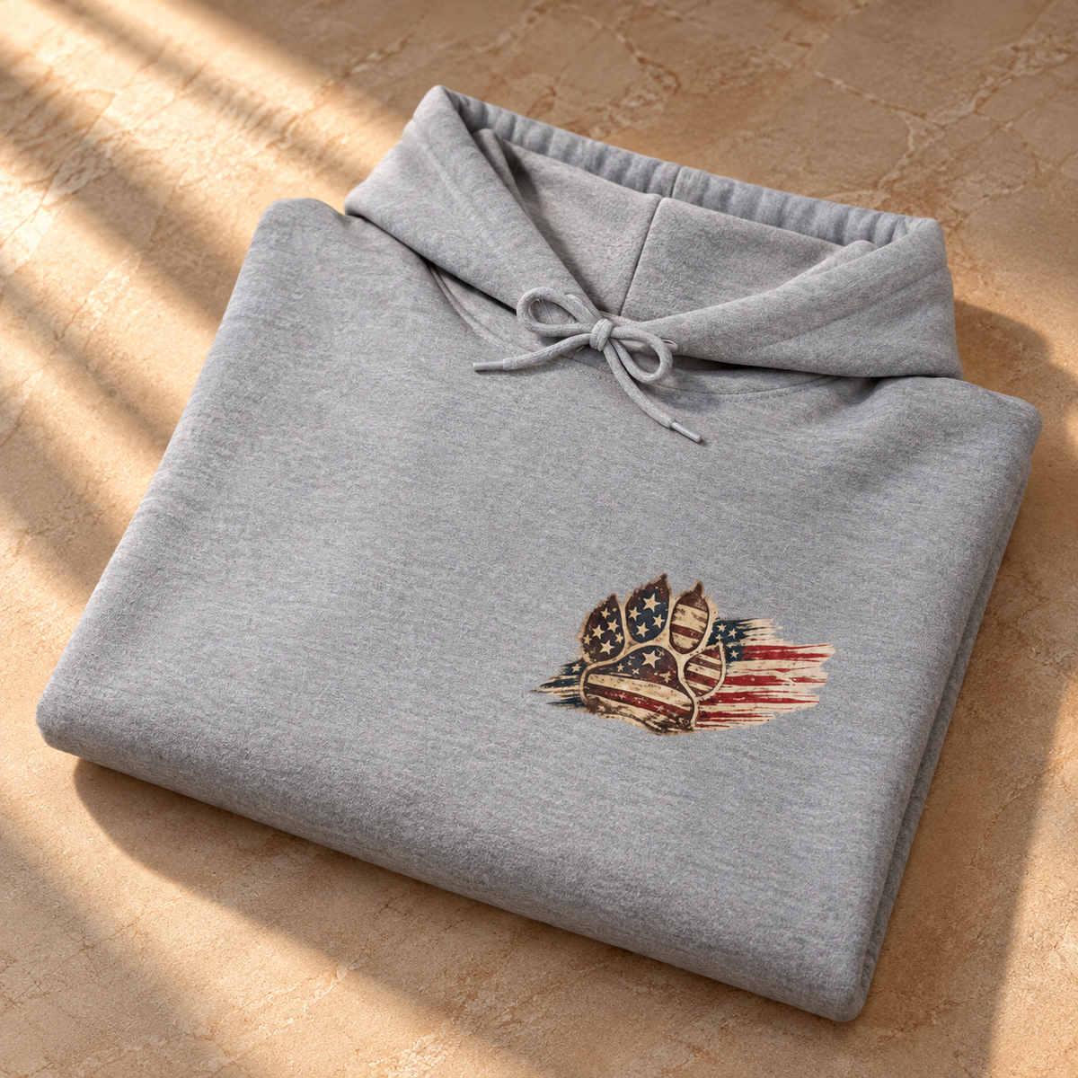A folded light gray heather hoodie with a small patriotic chest graphic featuring a dog silhouette and distressed American flag elements, displayed on a tan stone surface with diagonal window light shadows.