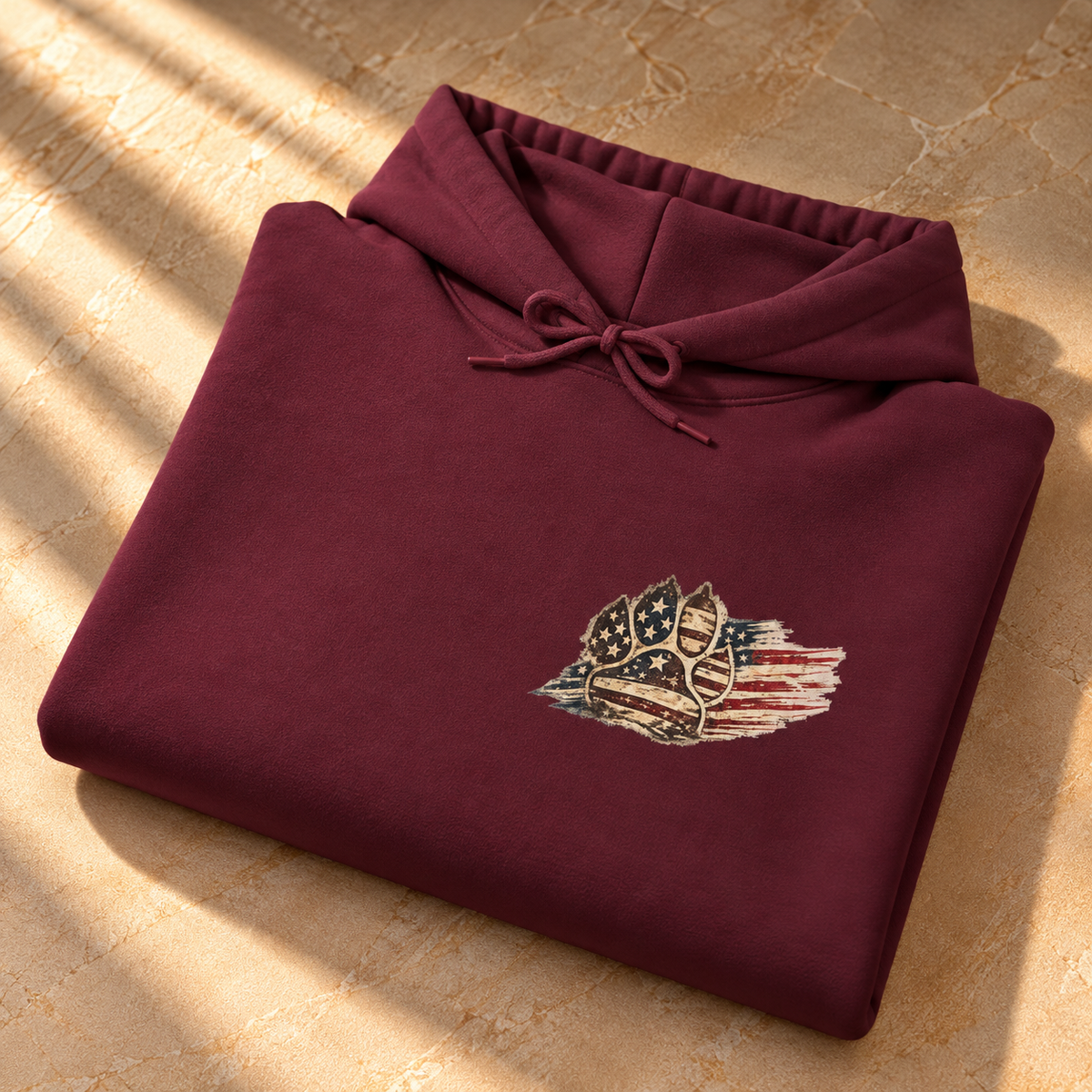 A folded maroon hoodie with a drawstring hood and a small patriotic chest graphic, placed on a textured beige surface with warm sunlight and shadows.
