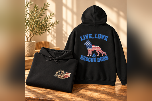 A black hoodie is shown folded on a wooden table next to its unfolded version, which features a large back graphic with the text 'LIVE, LOVE & RESCUE DOGS' in blue around a dog silhouette filled with the American flag. A smaller matching graphic is visible on the front pocket of the folded hoodie.